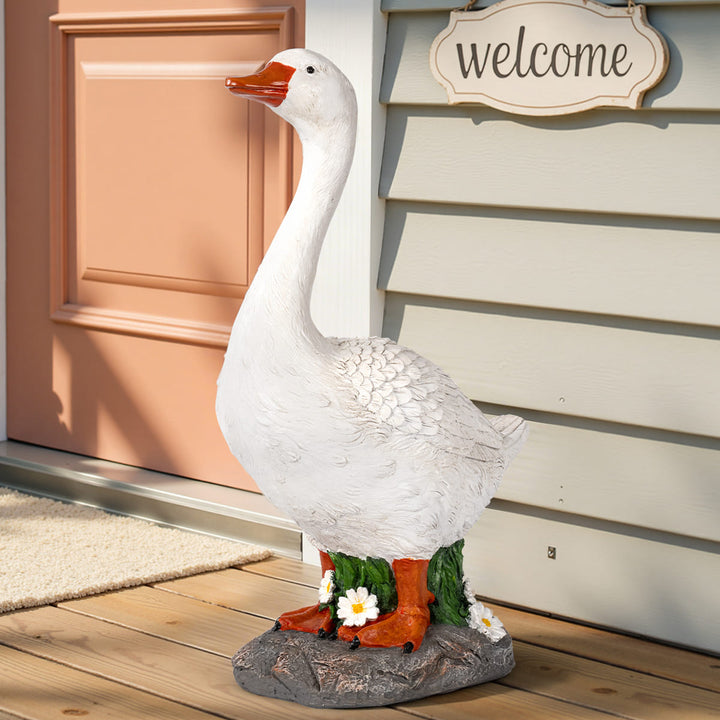 A white decorative goose statue with orange feet and beak stands on a wooden porch in front of a peach door and light gray siding. Small white flowers and green leaves emerge from the base, which is set on a gray rock-like pedestal.